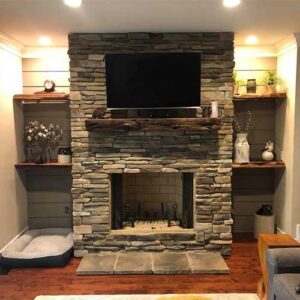 Wood Burning Fireplace with Stone Surround, hearth, and wooden mantel, shelving on each side with decorations - dog bed underneath left side shelving.