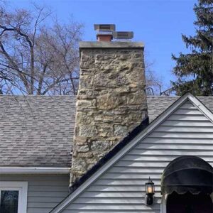 Restored stone masonry free of stains with new chimney crown and custom-fitted stainless steel chimney caps for both flues - Fall trees in the background with arched doorway on front of house.