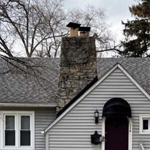 Dirty, stained stone chimney with double flue and deteriorating chimney caps - fall trees in the background with an arched doorway on the front.