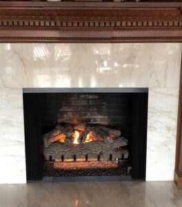 Fireplace with updated logs, black bricks and fire rocks white marble surround and chestnut wood mantel after facelift.