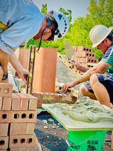 Techs doing masonry work rebuilding chimney - New bricks in the background and foreground building around the flue wearing safety helmets - Green trees in the background.